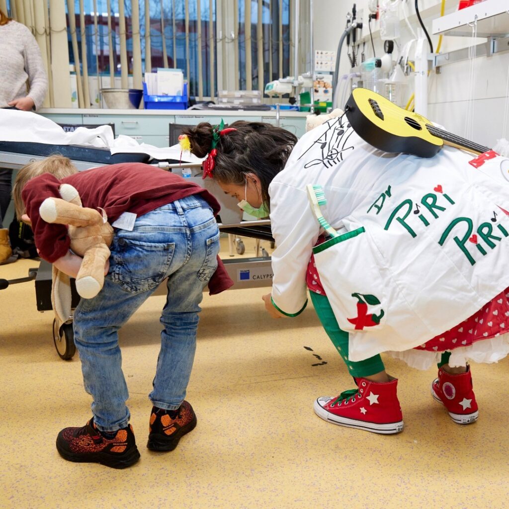 Un enfant se penche en tenant un animal en peluche tandis qu'une femme déguisée en clown avec une blouse blanche et des chaussures rouges imite les actions de l'enfant dans un contexte médical.