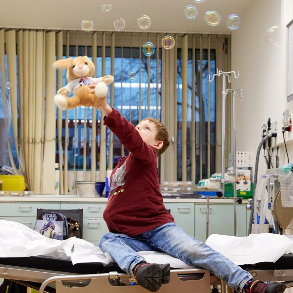 Un enfant portant une chemise marron et un jean est assis sur un lit d'hôpital, tendant la main vers un animal en peluche en direction de bulles flottantes dans une salle médicale.