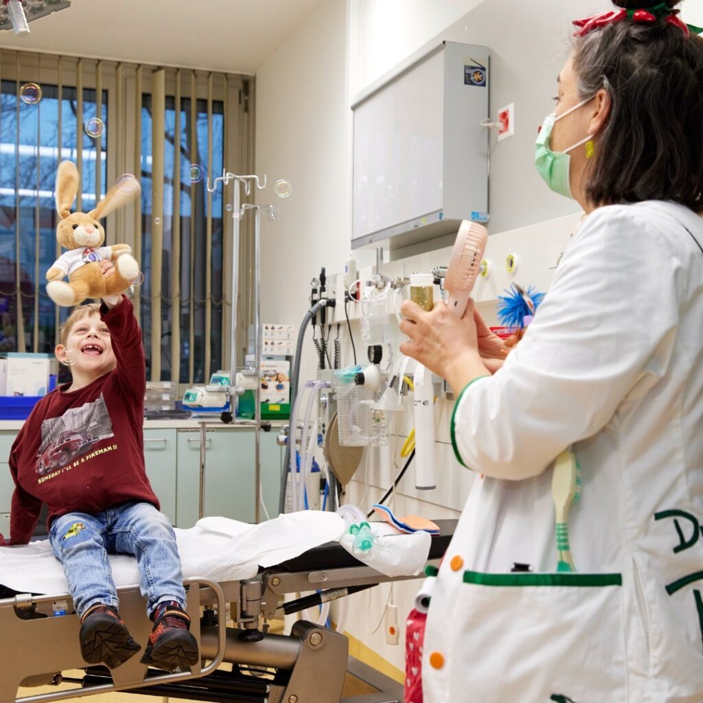 Un enfant sur un lit d'hôpital tend joyeusement la main vers un lapin en peluche tenu par un professionnel de la santé qui souffle des bulles.